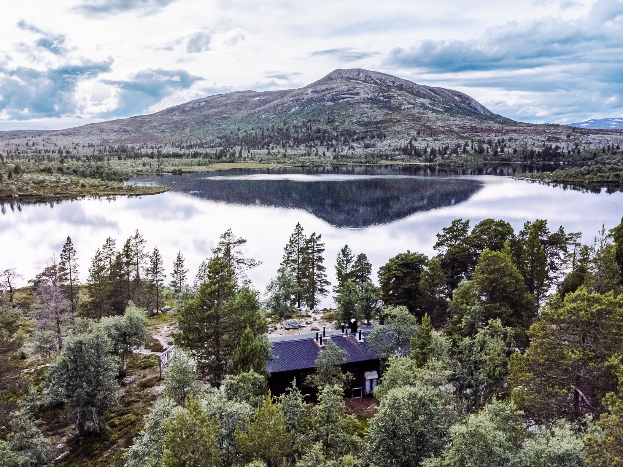 Fjällstuga vid sjö med fjäll och skog i bakgrunden.