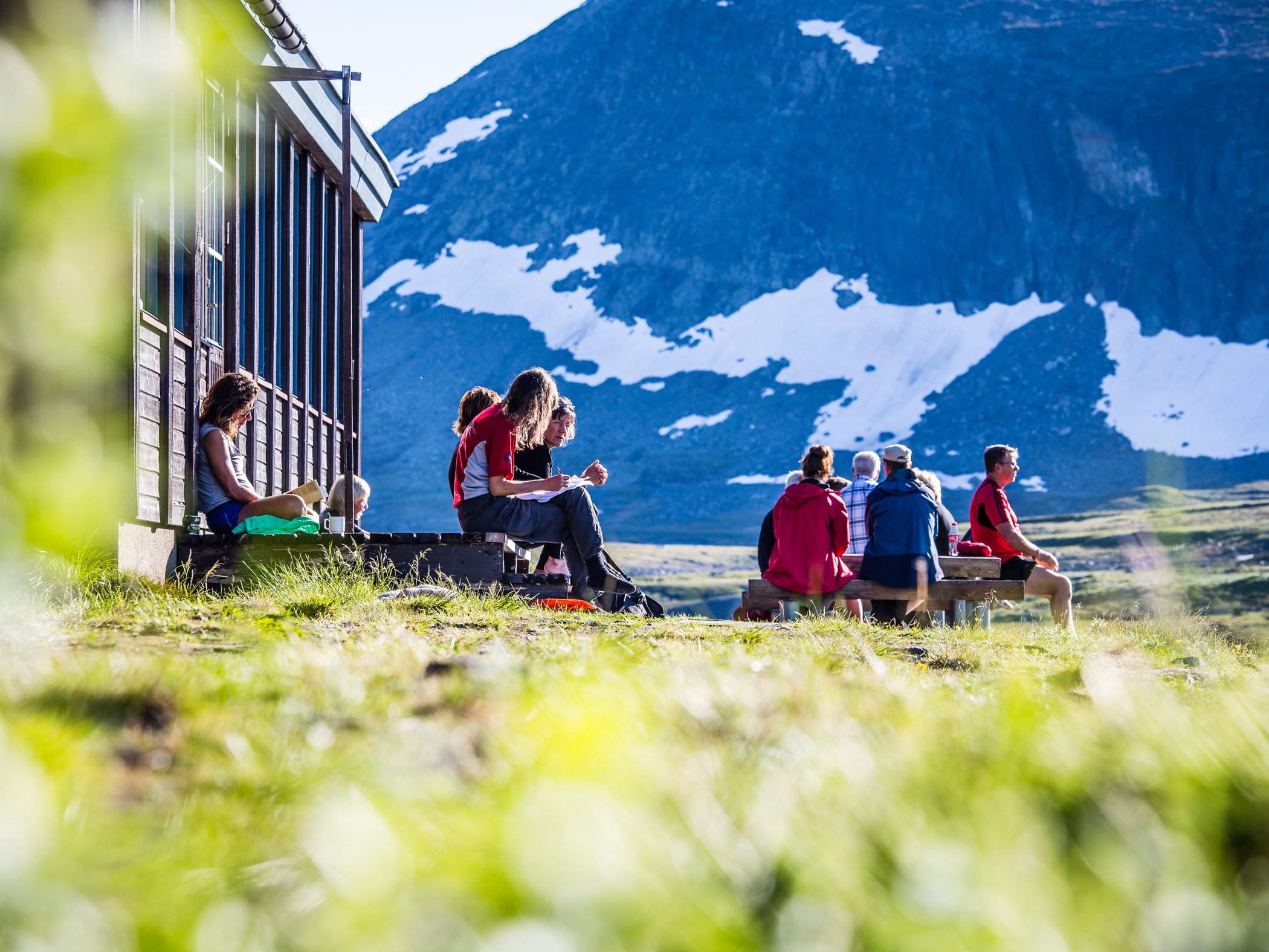 Personer sitter och vilar utanför fjällstation med utsikt över snöklädda fjäll.