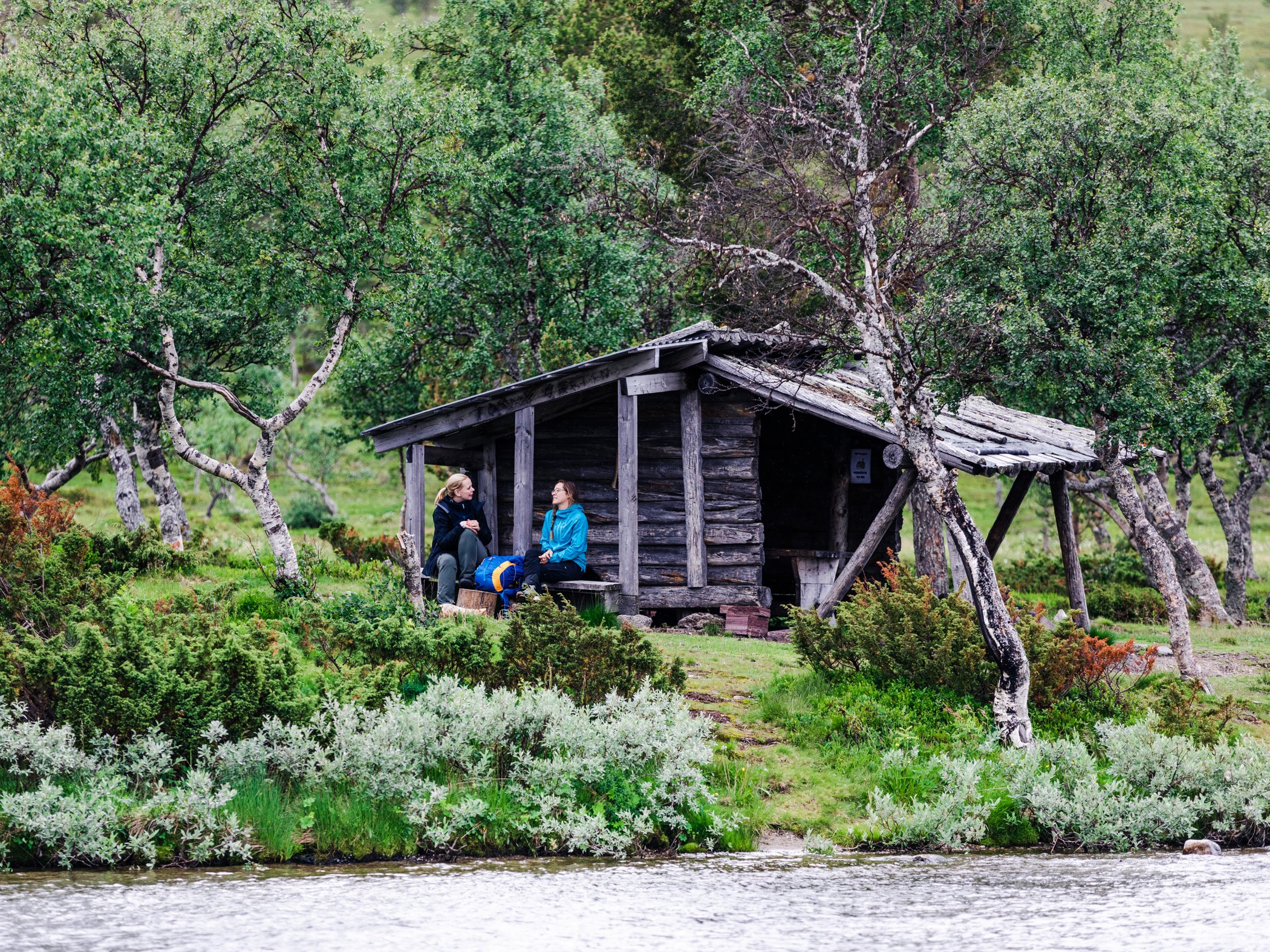 Två personer sitter vid en raststuga intill vatten i fjällmiljö.