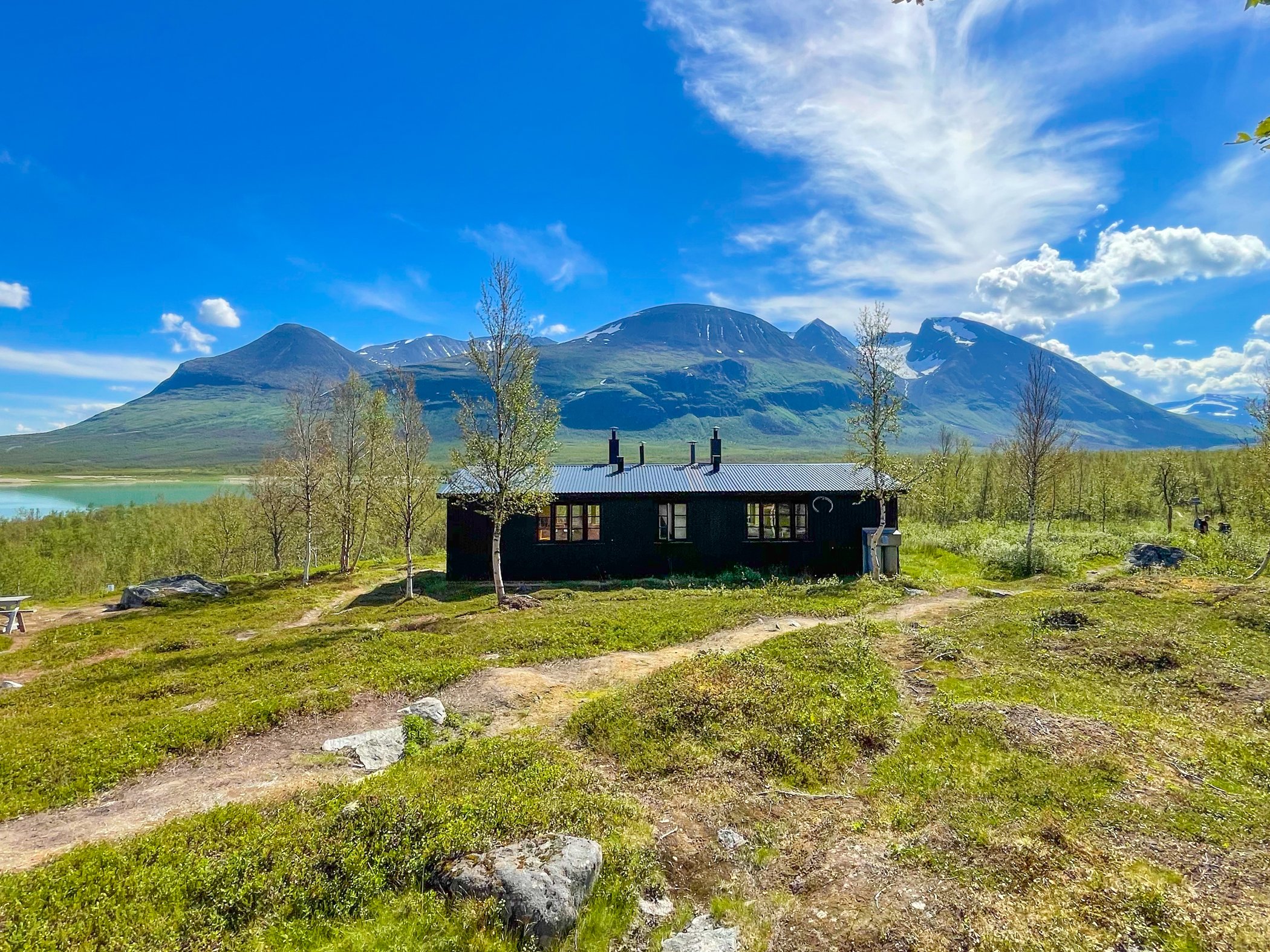 Fjällstuga med utsikt över grönt landskap och höga fjäll under blå himmel.