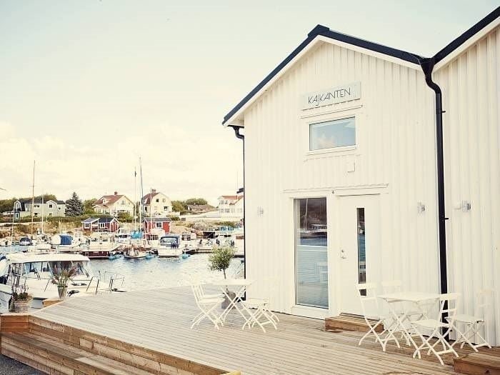Ljust sjöhus med terrass och uteservering vid hamnen, omgivet av båtar och kustbebyggelse.
