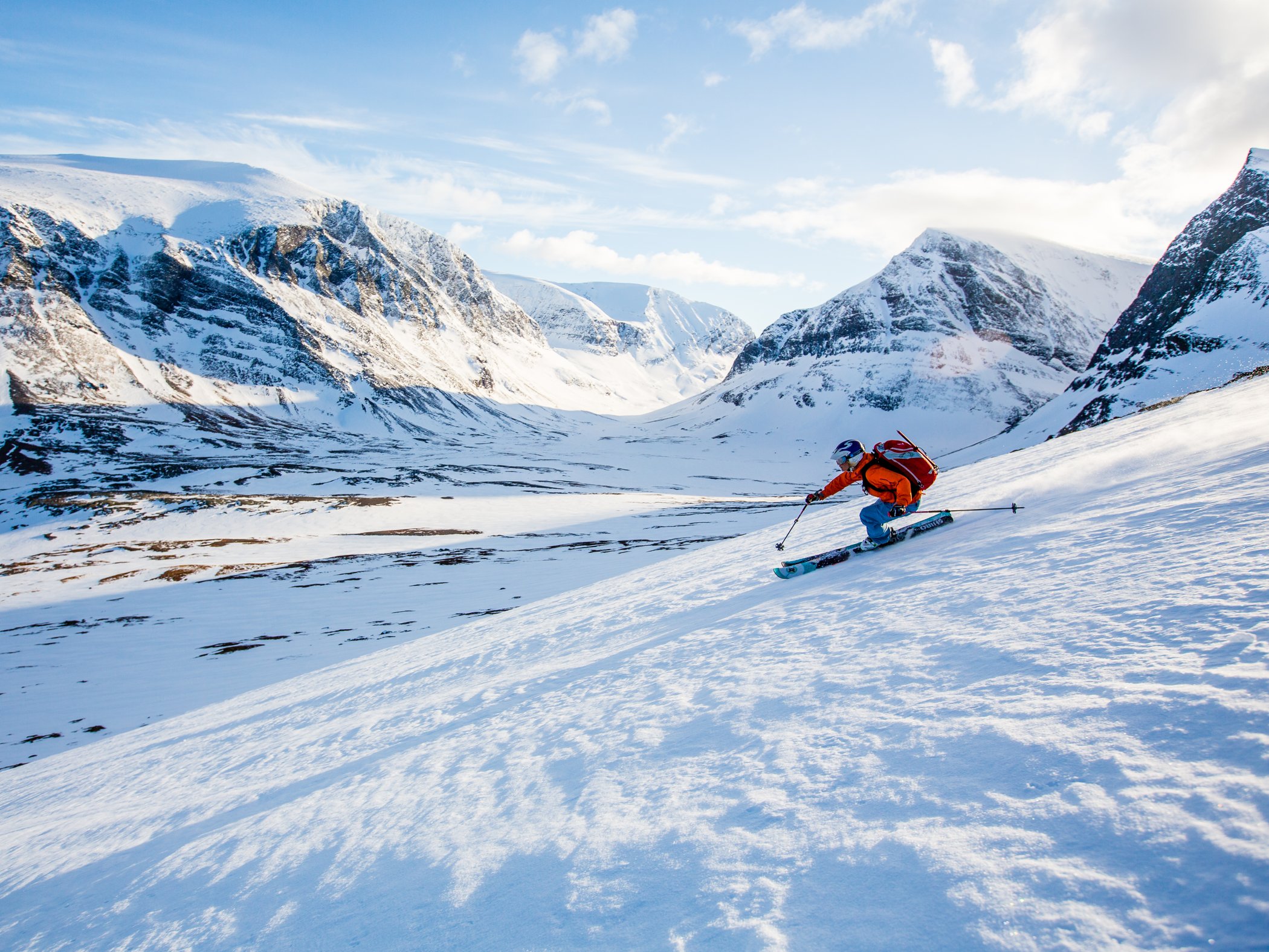 Person åker skidor nerför brant och snötackt fjällsida med dramatiska fjäll i bakgrunden.