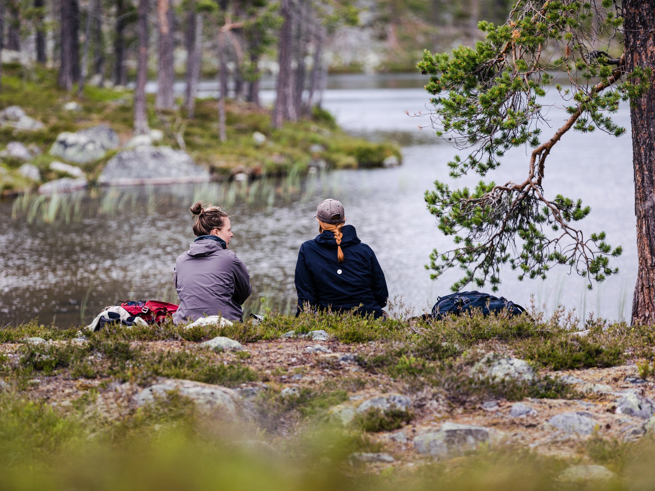 Två personer sitter vid en sjö i fjällmiljö och tar paus.