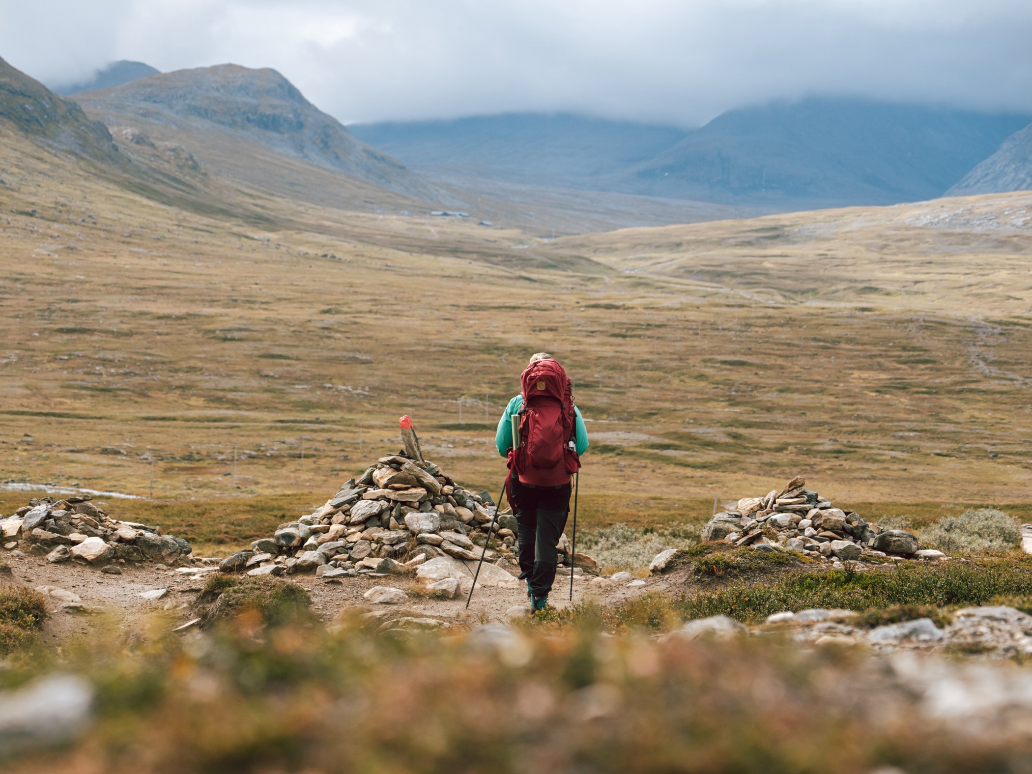 Vandrare med stor ryggsäck går på fjällstig genom öppen dalgång mellan fjäll.