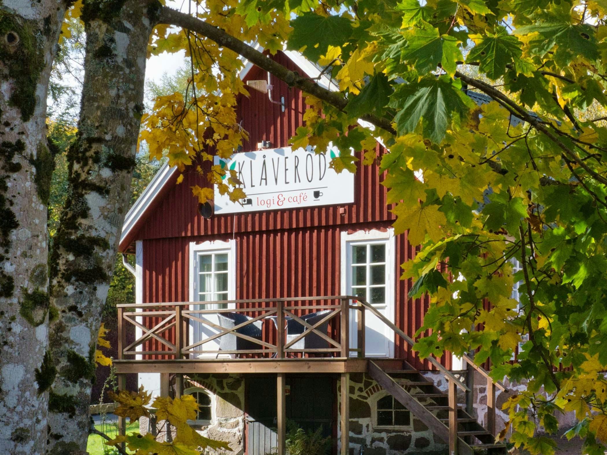 Röd träbyggnad med skylt för Klåveröd logi & café, inramad av gulgröna höstlöv och träd i förgrunden.