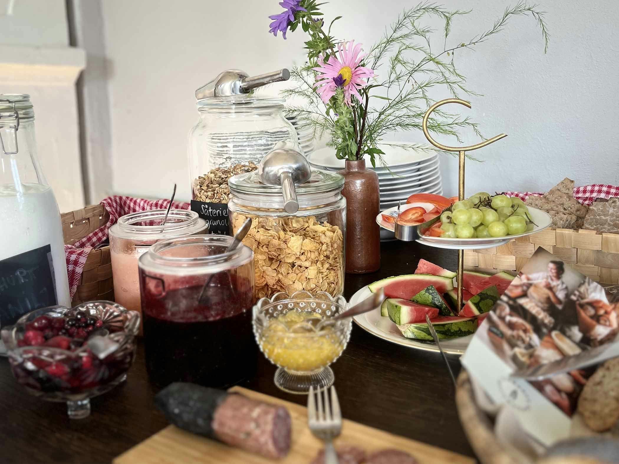 Frukostbuffé med müsli, sylt, bär, vattenmelon, vindruvor, bröd och pålägg uppdukat på ett bord med färska blommor.