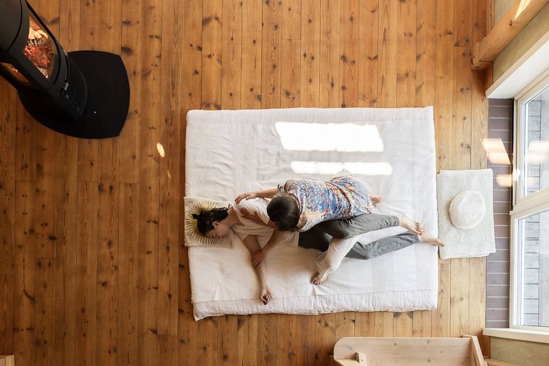 Yogamassagebehandling sedd ovanifrån där en person får assisterad stretch på en madrass i en träinredd lokal.