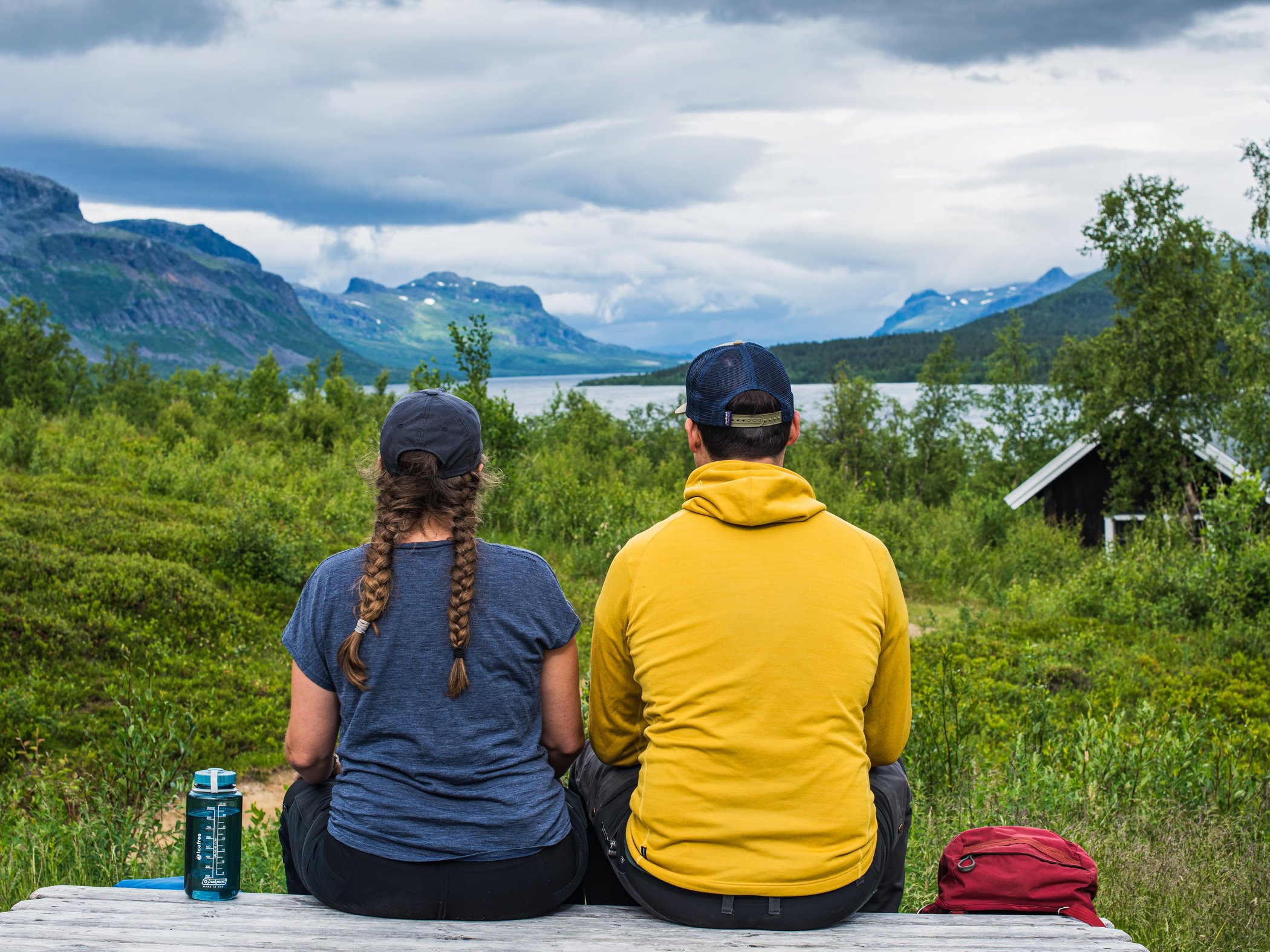 Par sitter på bänk och ser ut över fjällsjö och gröna fjäll.