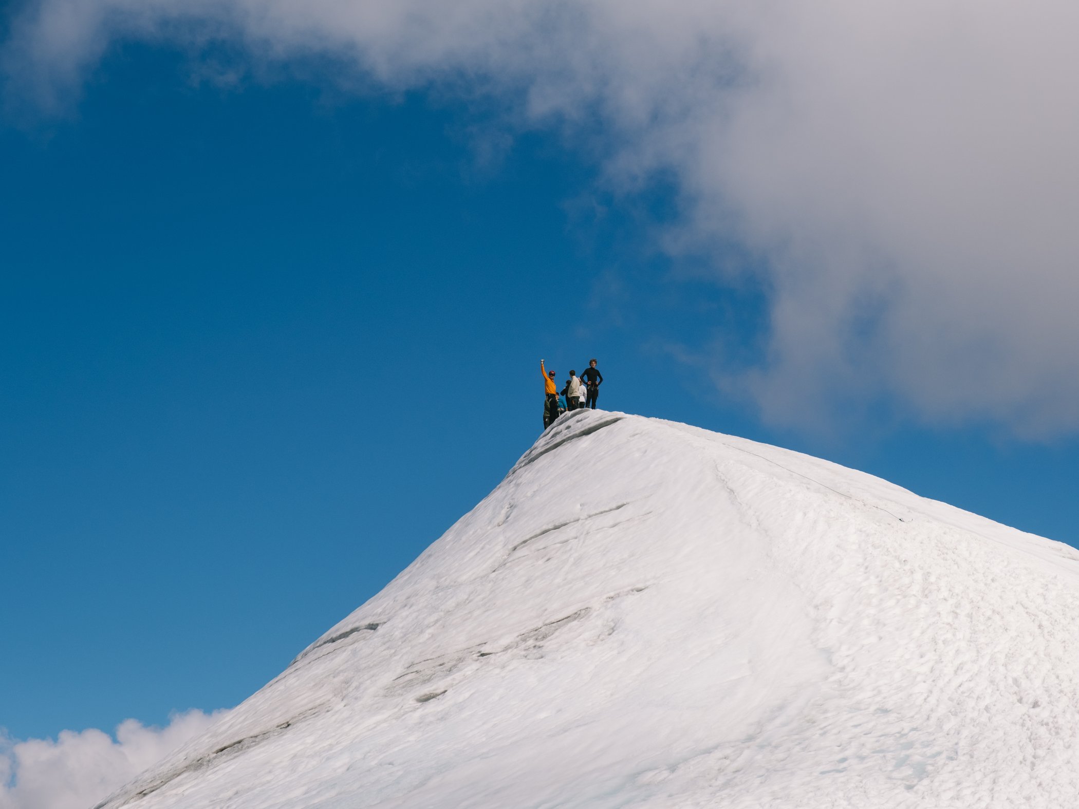Tre personer står på en snötäckt fjälltopp under blå himmel.
