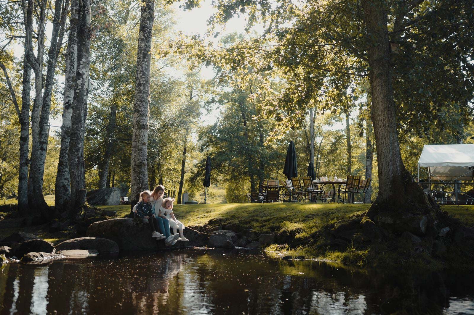 Personer sitter vid en liten bäck i en lummig park med träd och utemöbler.