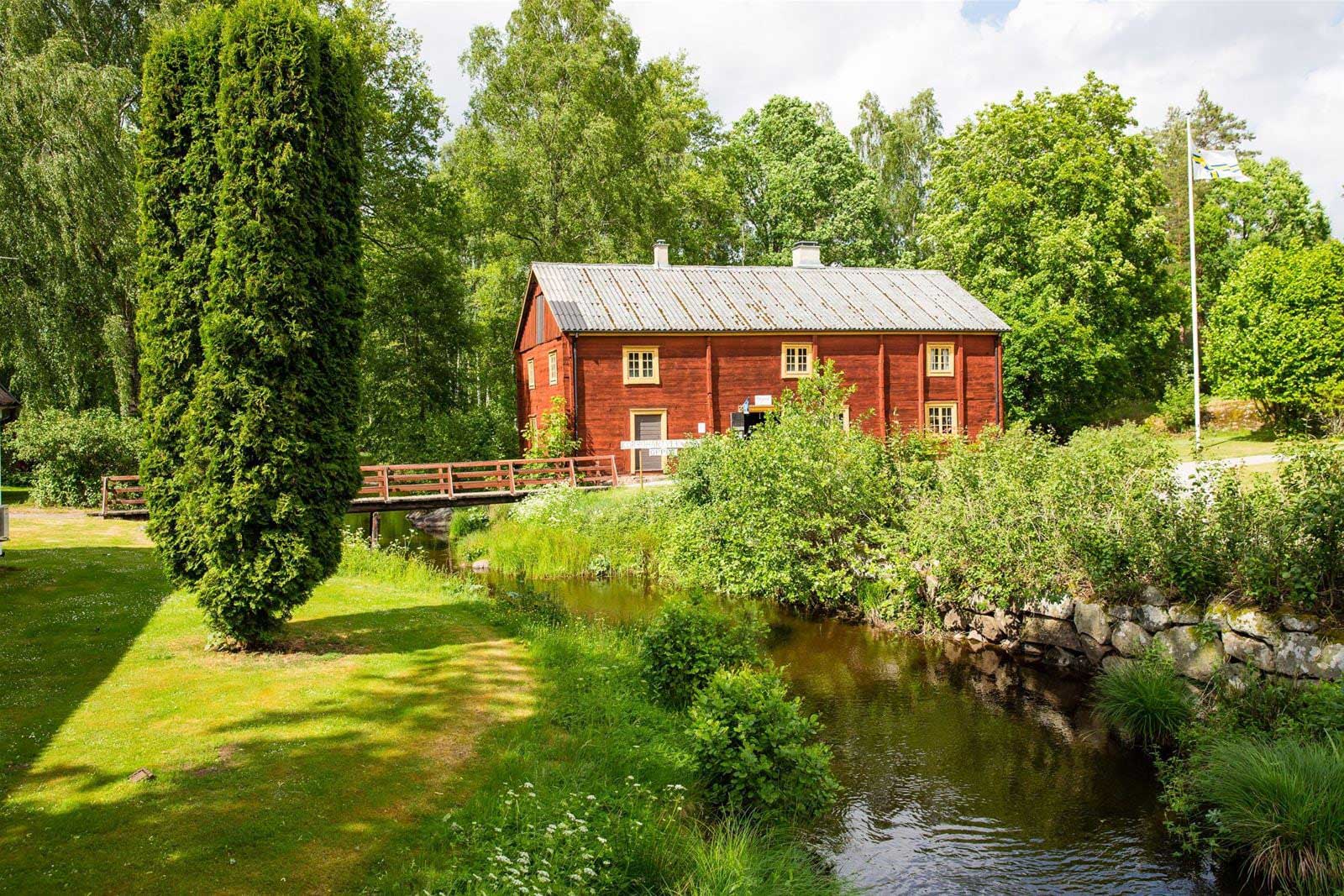Röd träbyggnad vid en bäck med grönskande omgivning och liten bro.