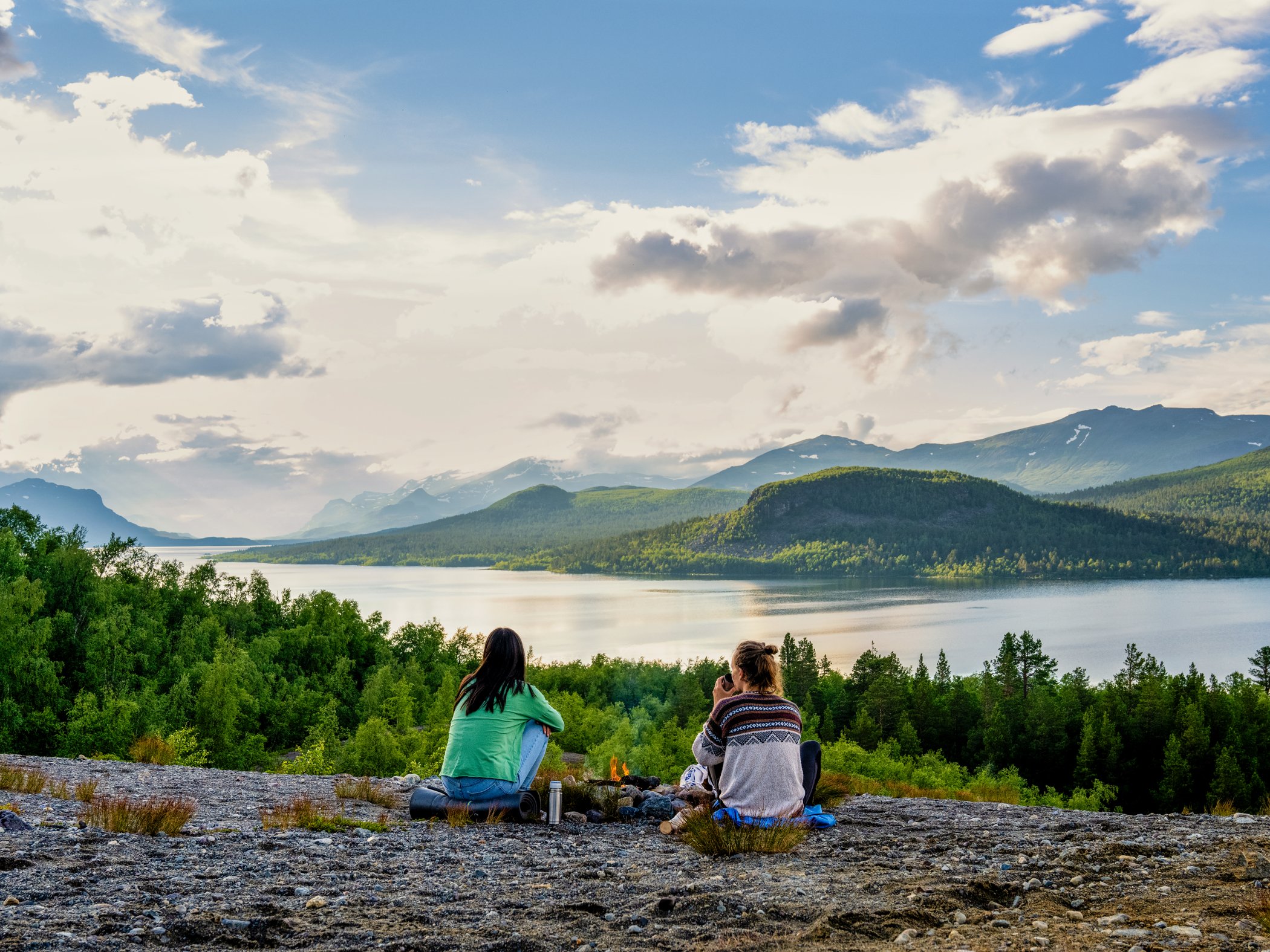 Två personer sitter vid utsiktsplats med sjö och fjäll i kvällsljus.