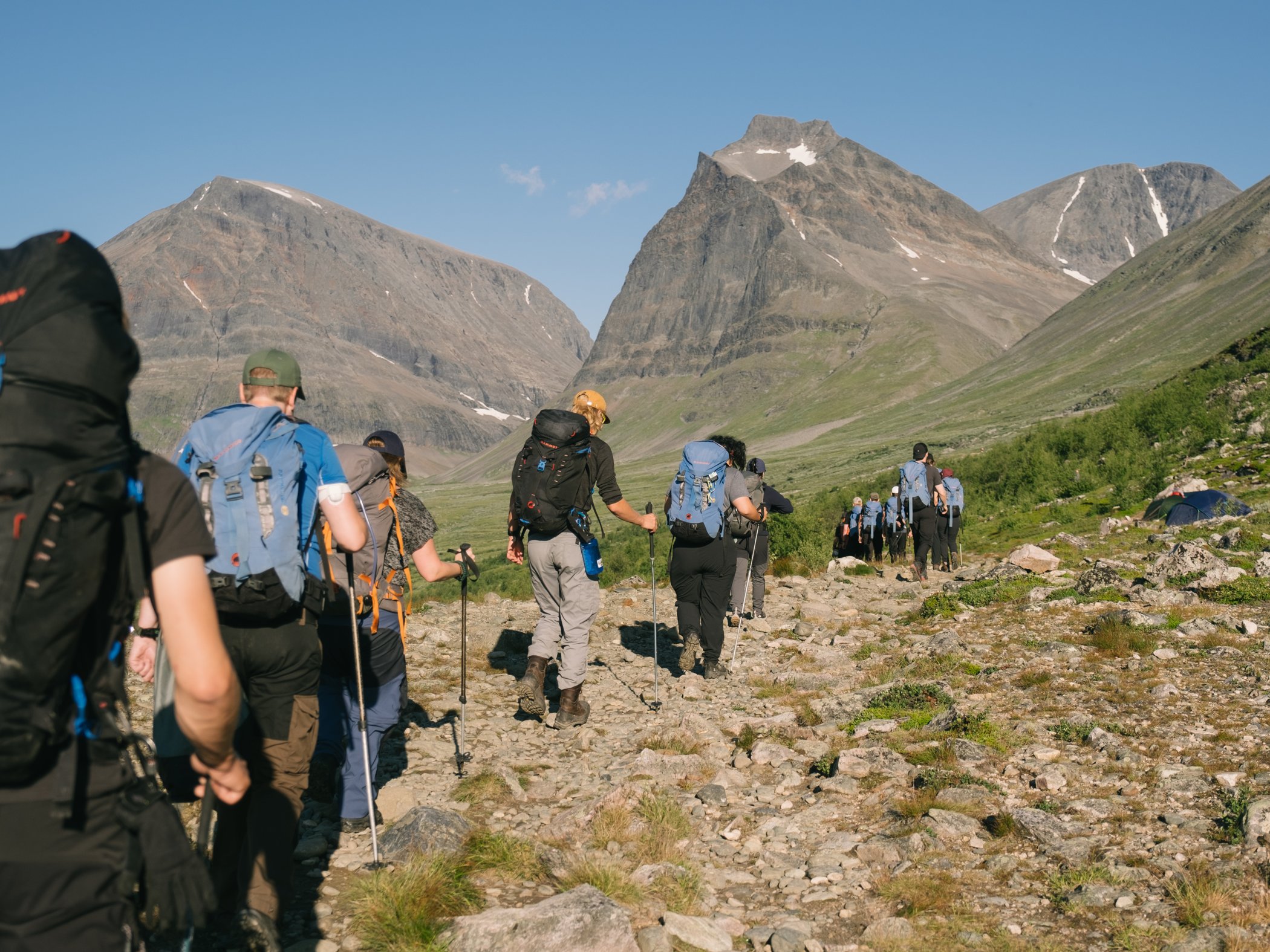 Grupp av vandrare på led genom fjällandskap med höga berg i bakgrunden.