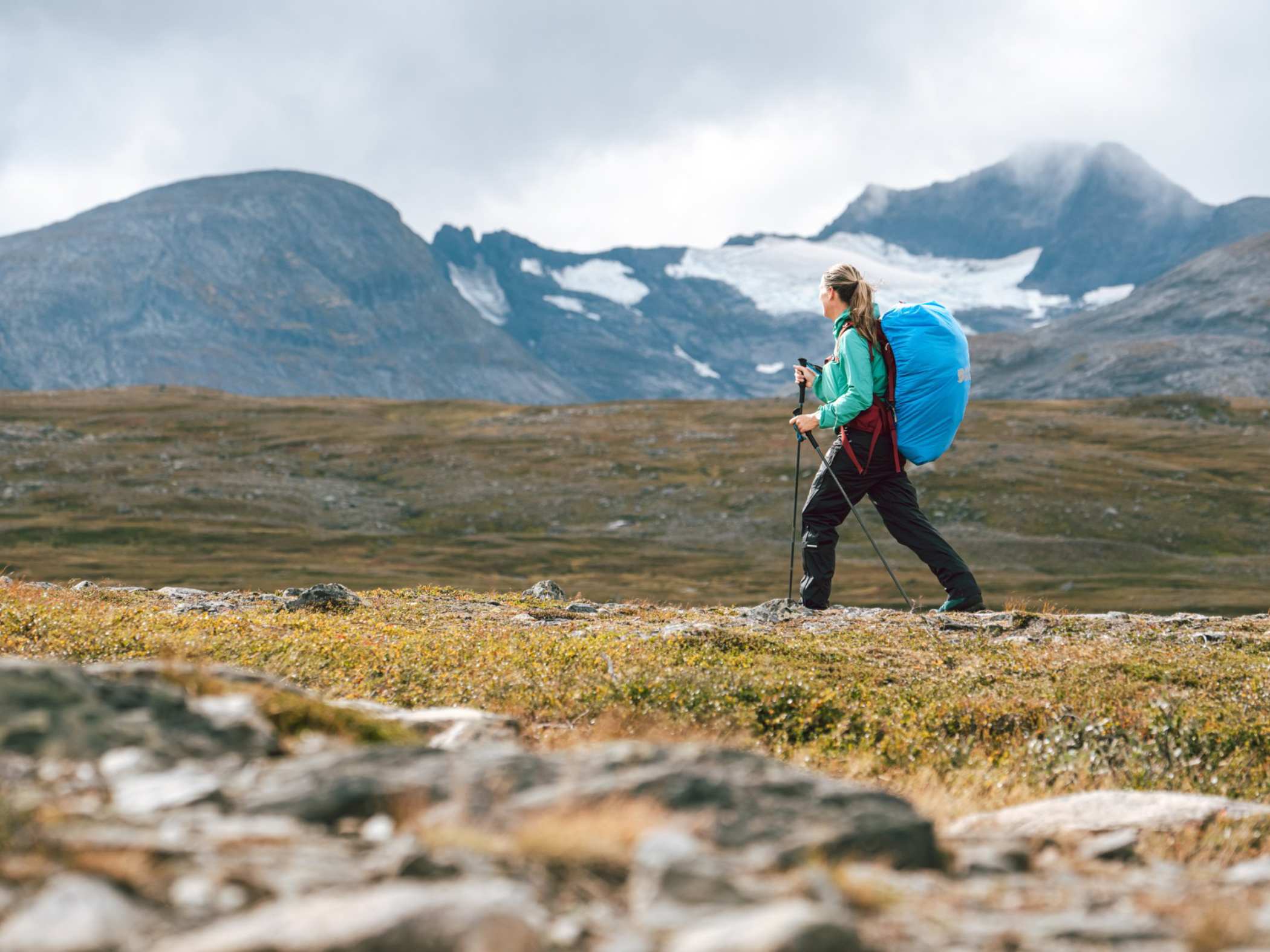 Vandrare med stor ryggsäck och vandringsstavar på kalfjäll med glaciär och fjälltoppar i bakgrunden.