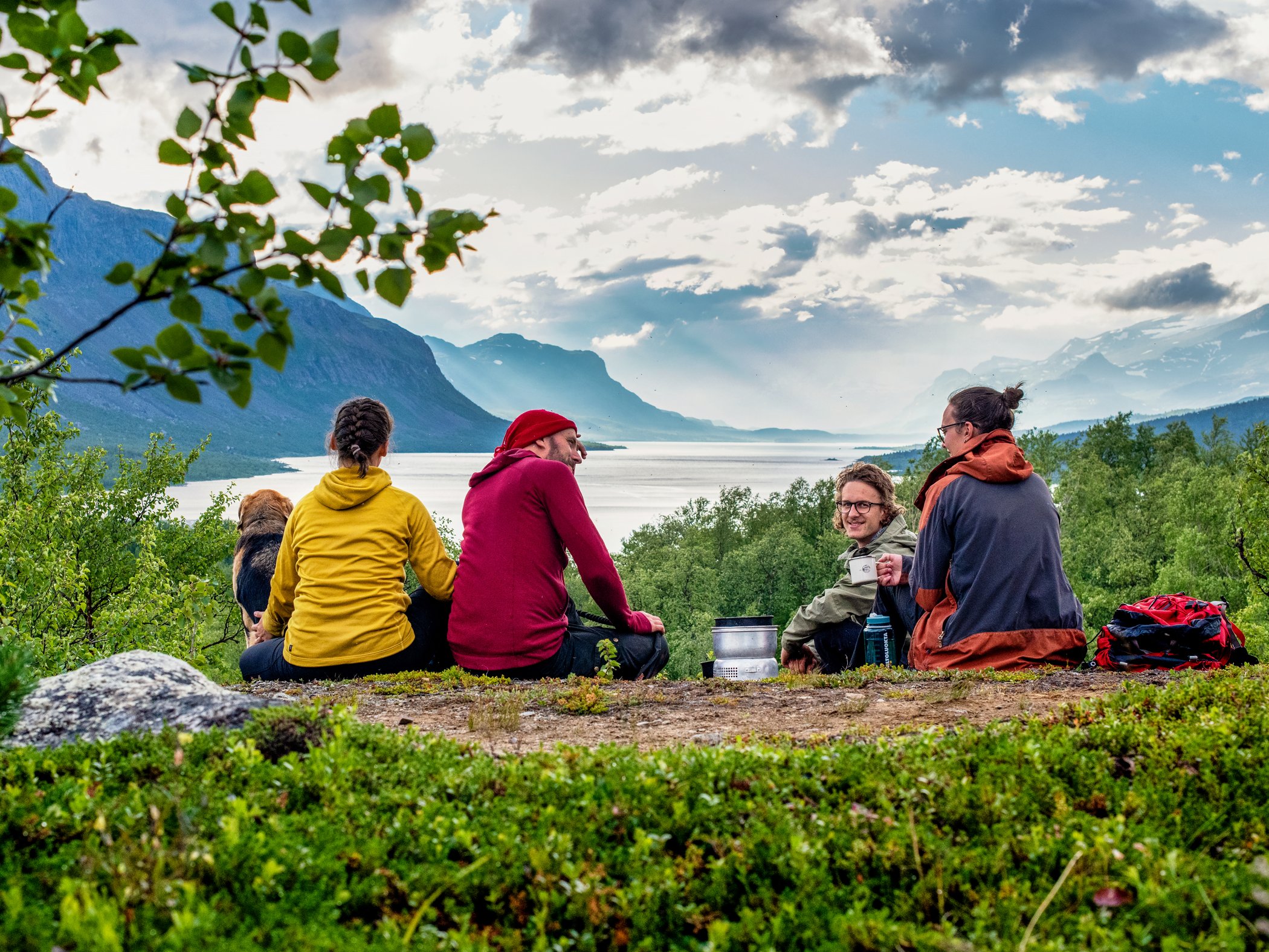 Vandrare och hund pausar med utsikt över fjällsjö och dramatiska moln.