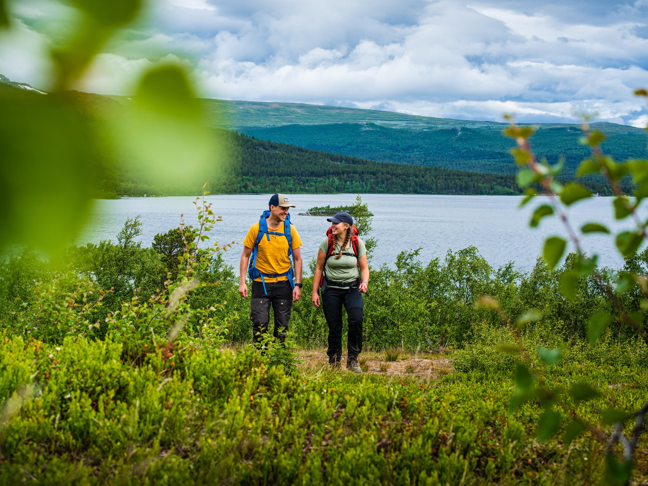 Två vandrare med ryggsäckar går genom grönskande landskap vid sjö och fjäll.
