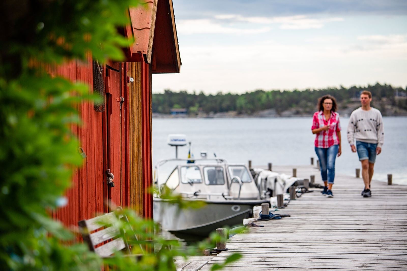 Två personer promenerar på en brygga vid vattnet intill röda sjöbodar och en förtöjd båt.