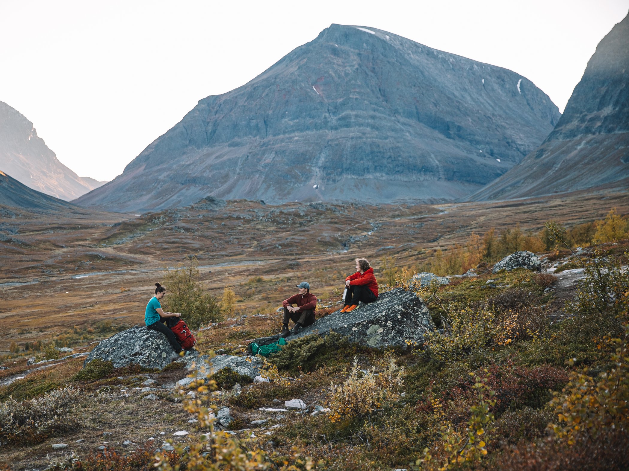 Tre vandrare sitter på stenar i fjällandskap med höstfärger och berg i bakgrunden.