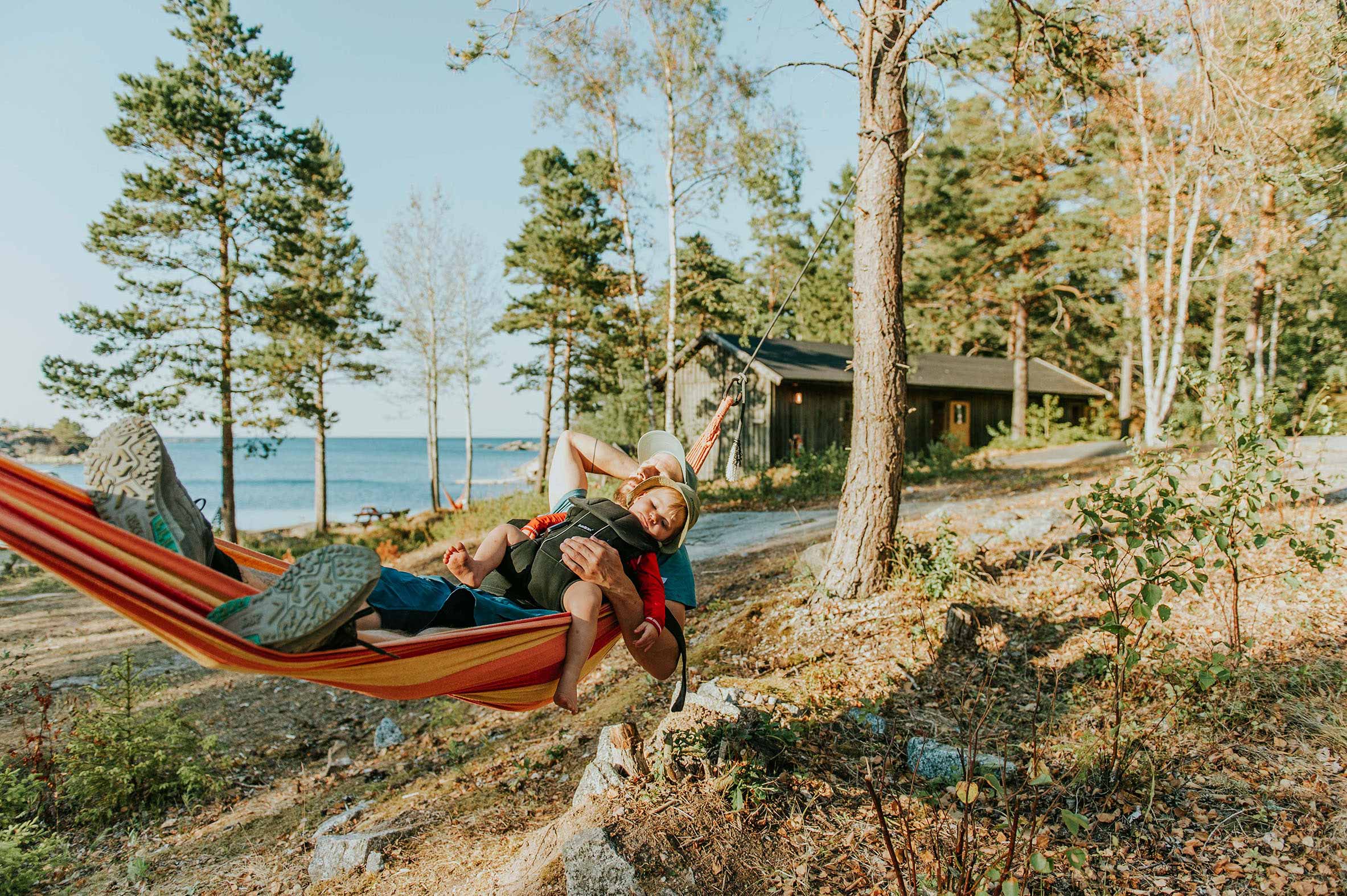 Person vilar i en hängmatta med ett barn vid havet intill en stuga i skogen.