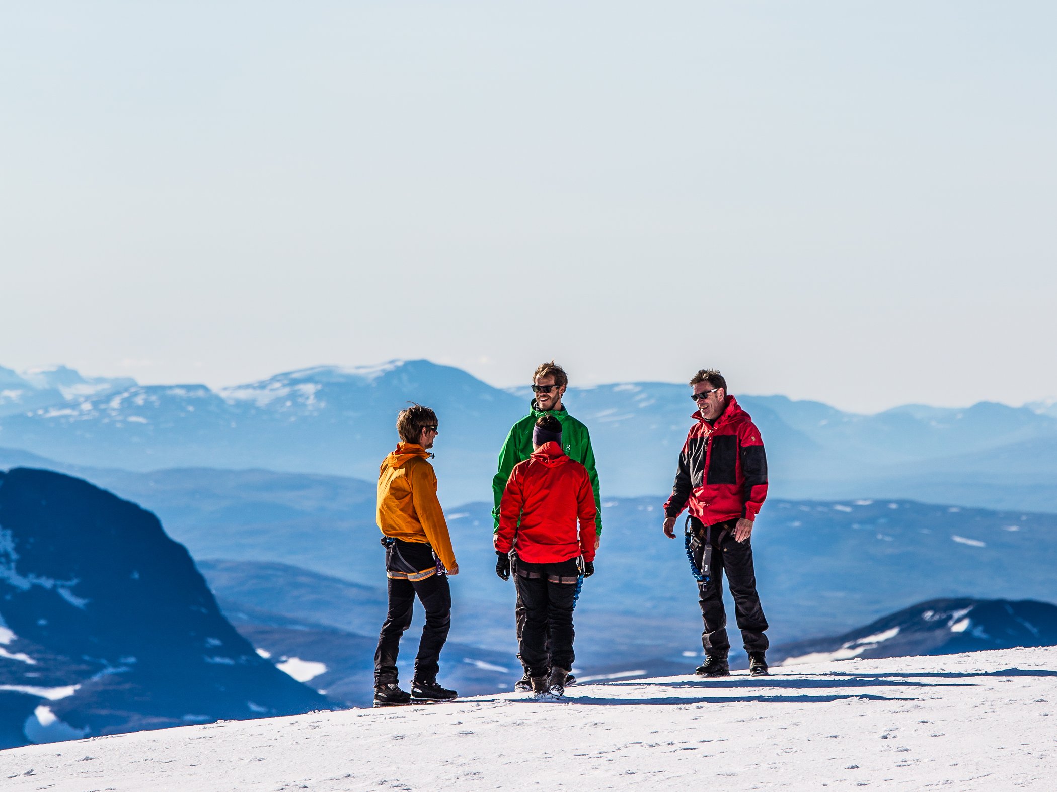 Fyra personer står på snötäckt fjäll med bergslandskap i bakgrunden.