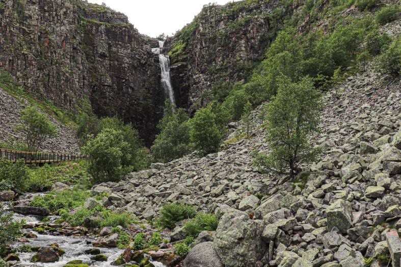 Vattenfall som störtar ner mellan branta klippväggar omgivet av stenblocksterräng och fjällbjörkar.
