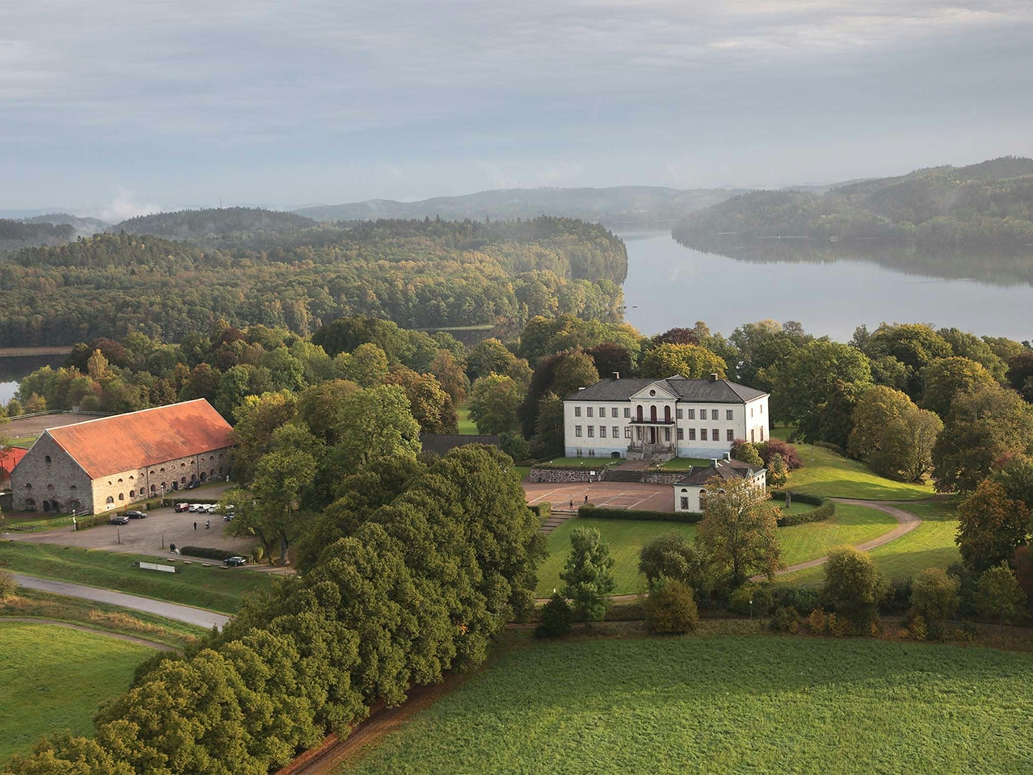 Flygbild över en herrgård med omgivande parklandskap, skog och sjö i ett böljande kulturlandskap.