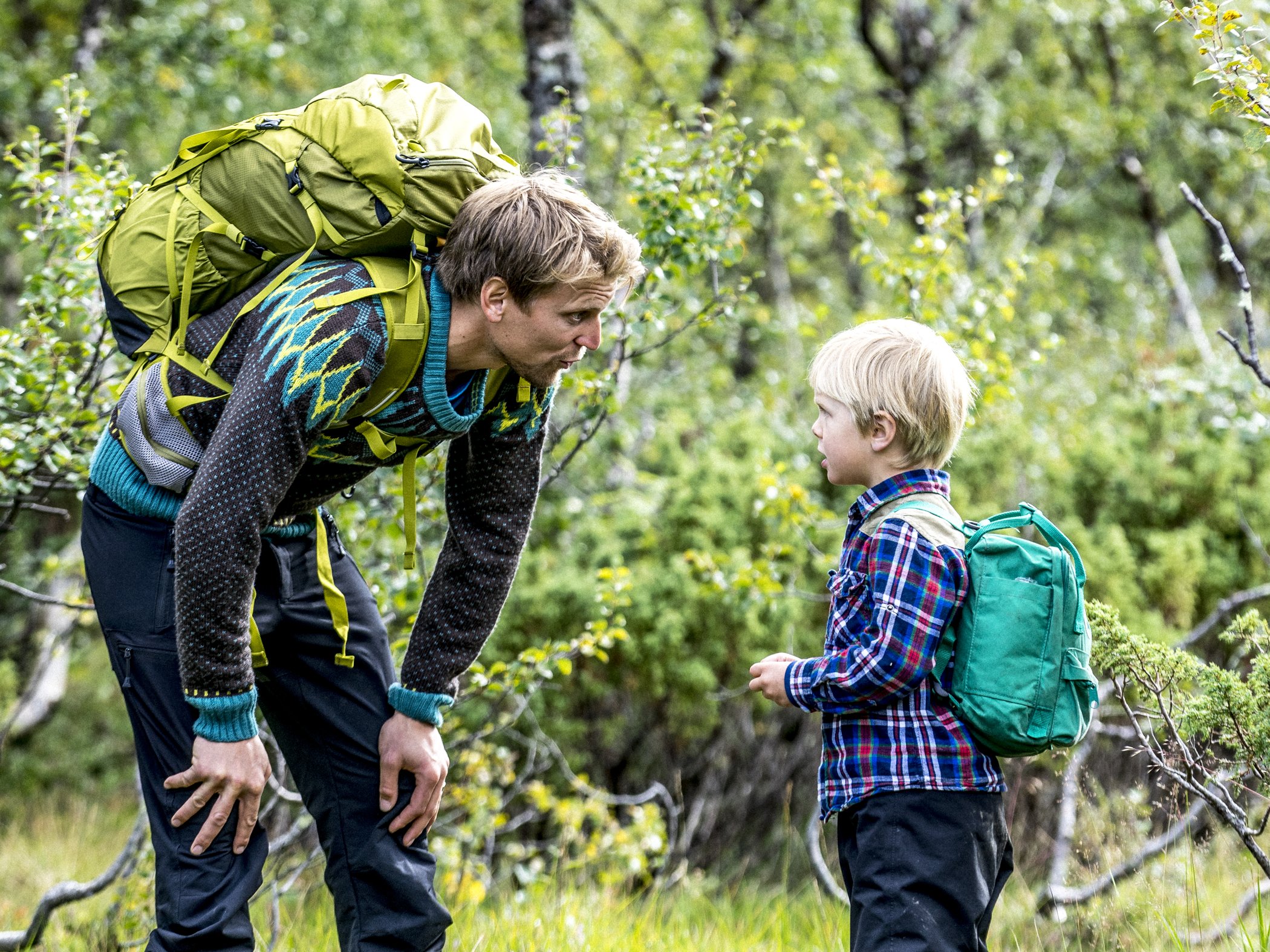 Vuxen och barn med ryggsäckar på vandring i fjällskog.