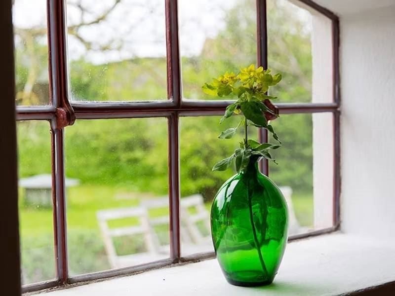 Grön glasflaska med gula blommor på en fönsterbräda med utsikt mot en grönskande trädgård.