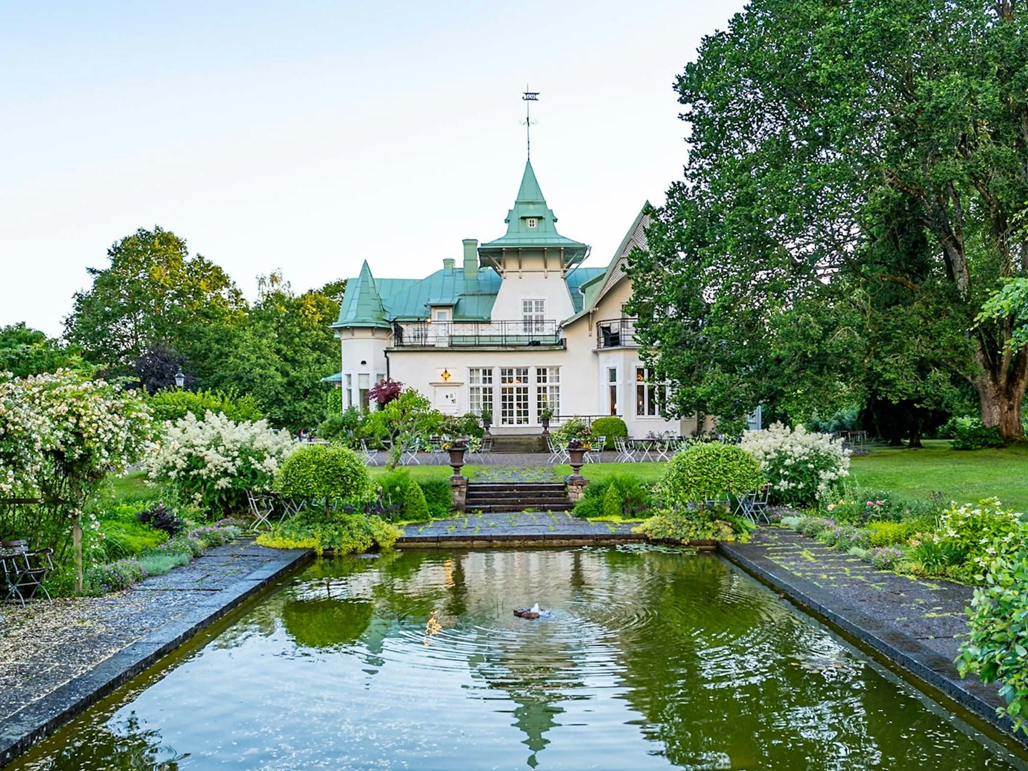 Herrgård omgiven av välskött trädgård med spegeldamm, blommande buskar och stora lövträd.