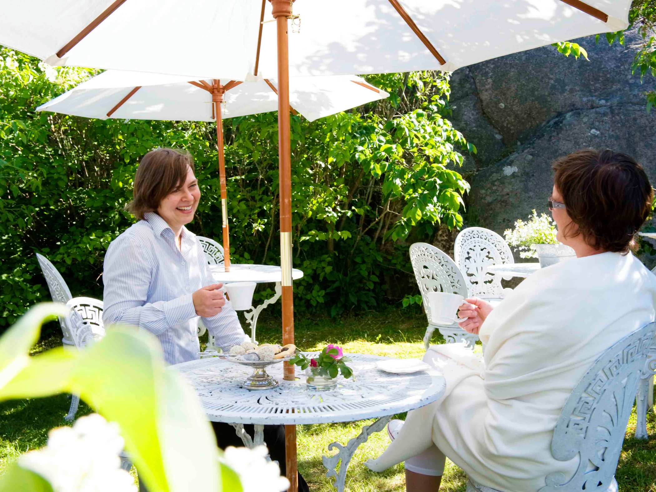 Två kvinnor som sitter under vita parasoll och dricker kaffe i grönskan. 