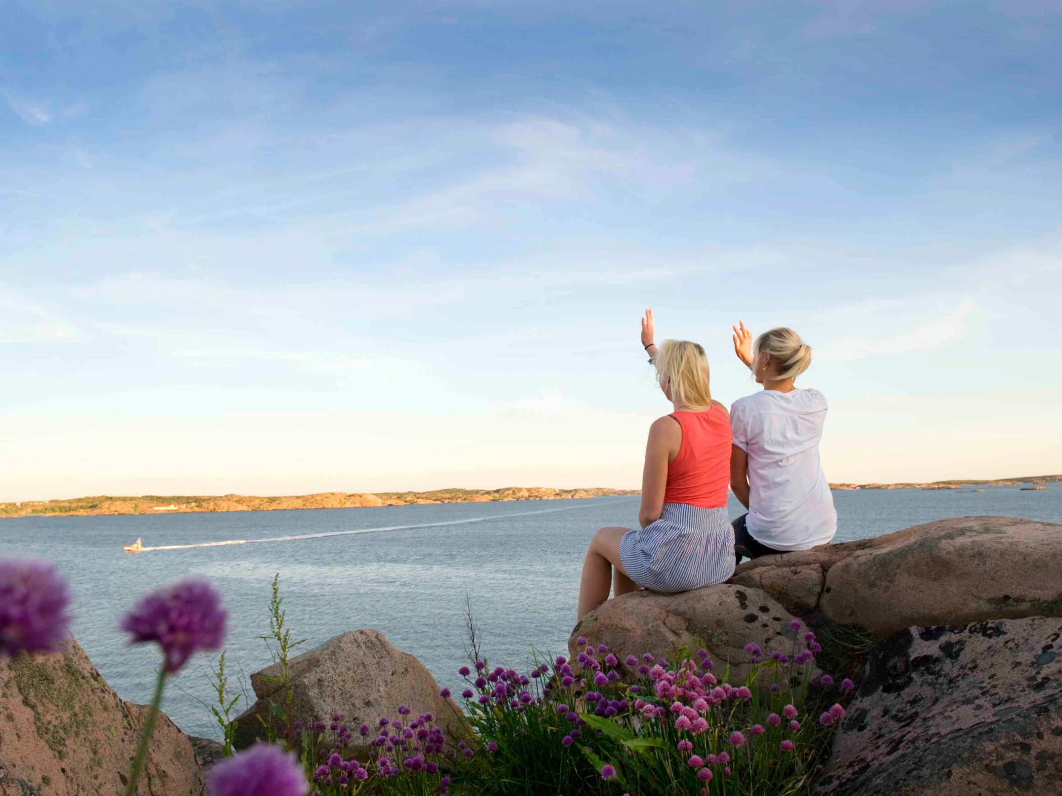 Två kvinnor som sitter på en klippa och blickar ut över ett soligt hav. 
