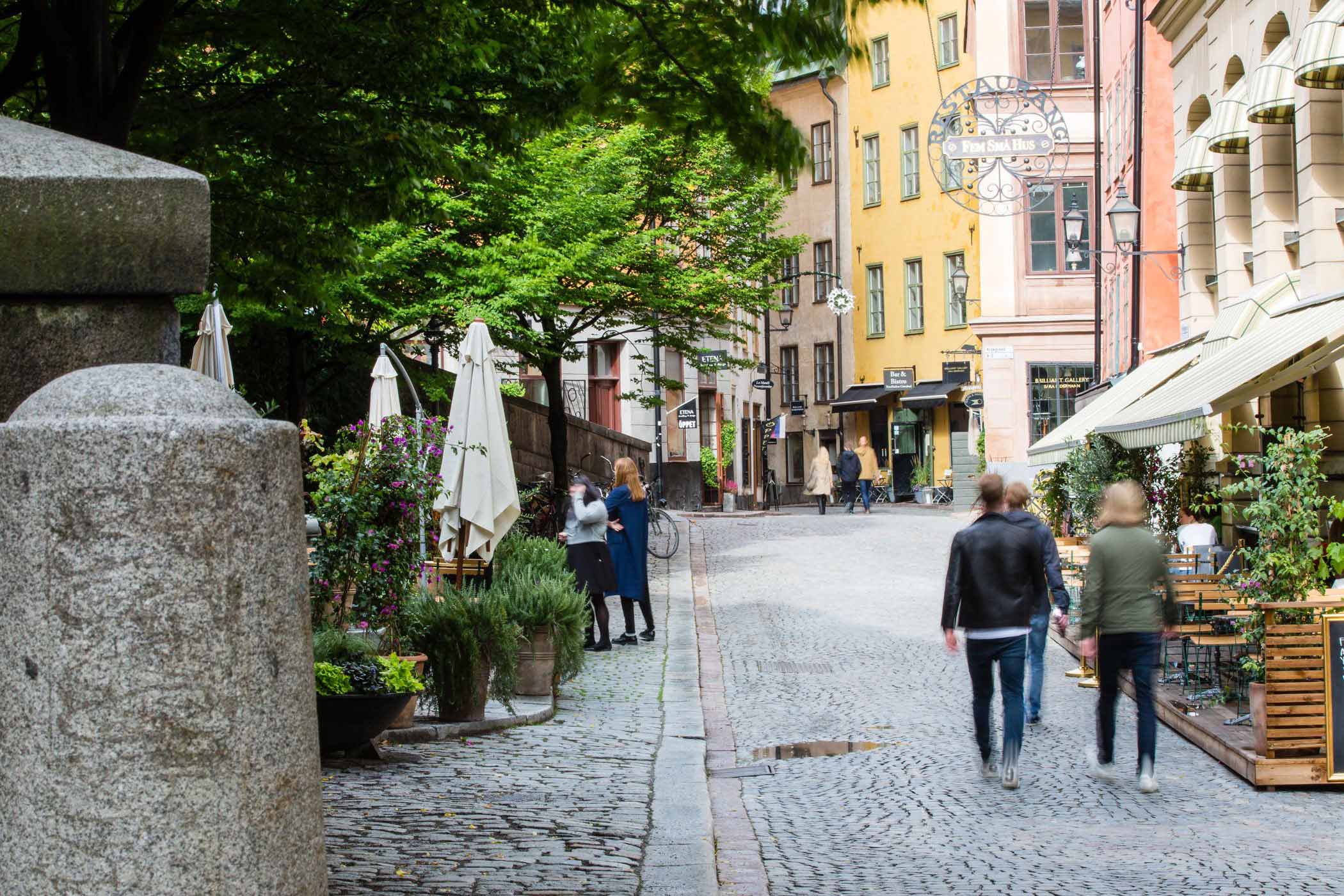 Människor promenerar på en kullerstensgata i Gamla stan med färgglada hus och uteserveringar.