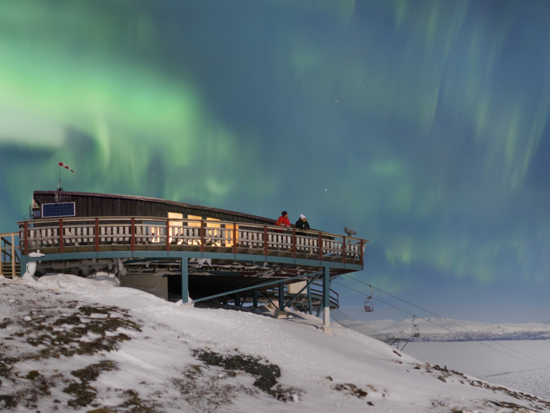 Två personer på terrassen vid STF Abisko Fjällstation medan norrsken lyser över det snötäckta fjällandskapet.