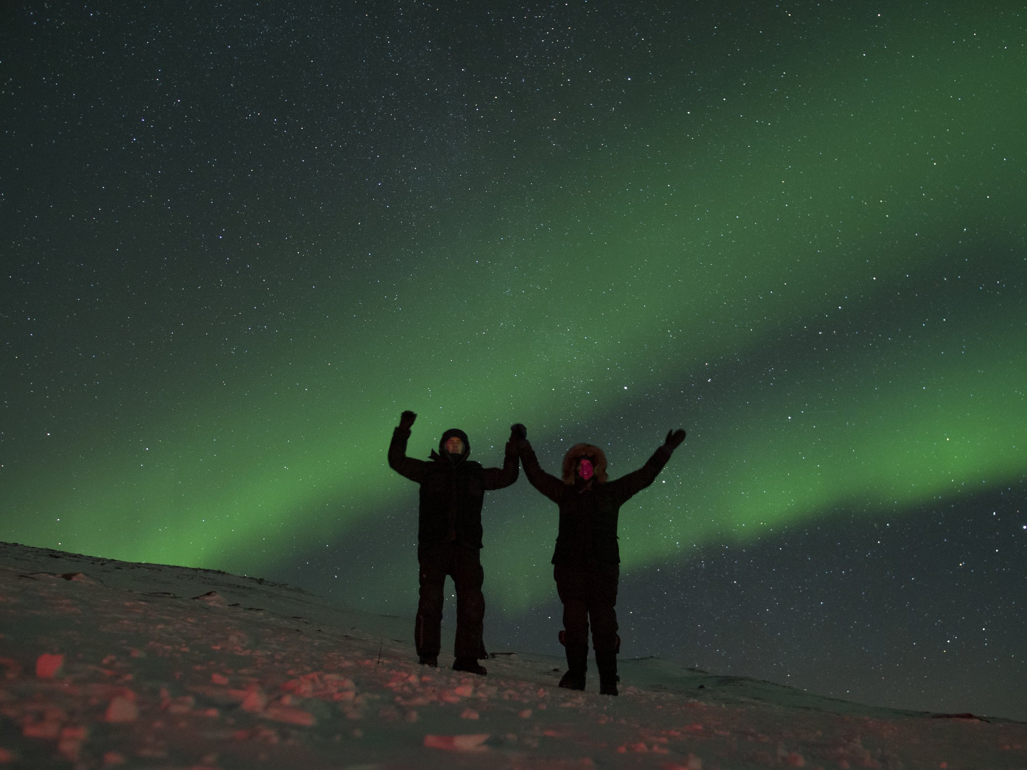 Två personer står i snön och ser norrsken dansa över den stjärnklara himlen i Abisko.