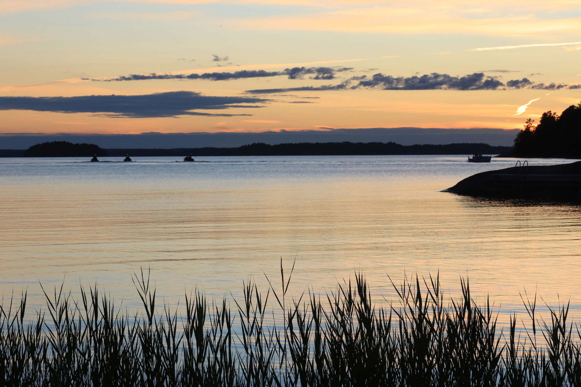 Solnedgång över lugnt vatten i skärgården med siluetter av vass och båtar.