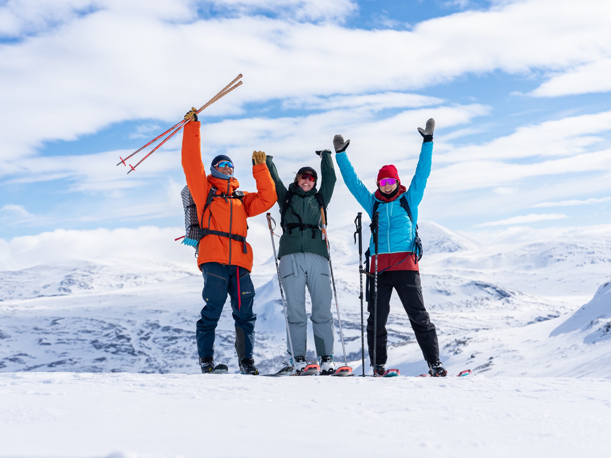 Tre personer firar på fjället efter en topptur med snötäckta berg i bakgrunden.