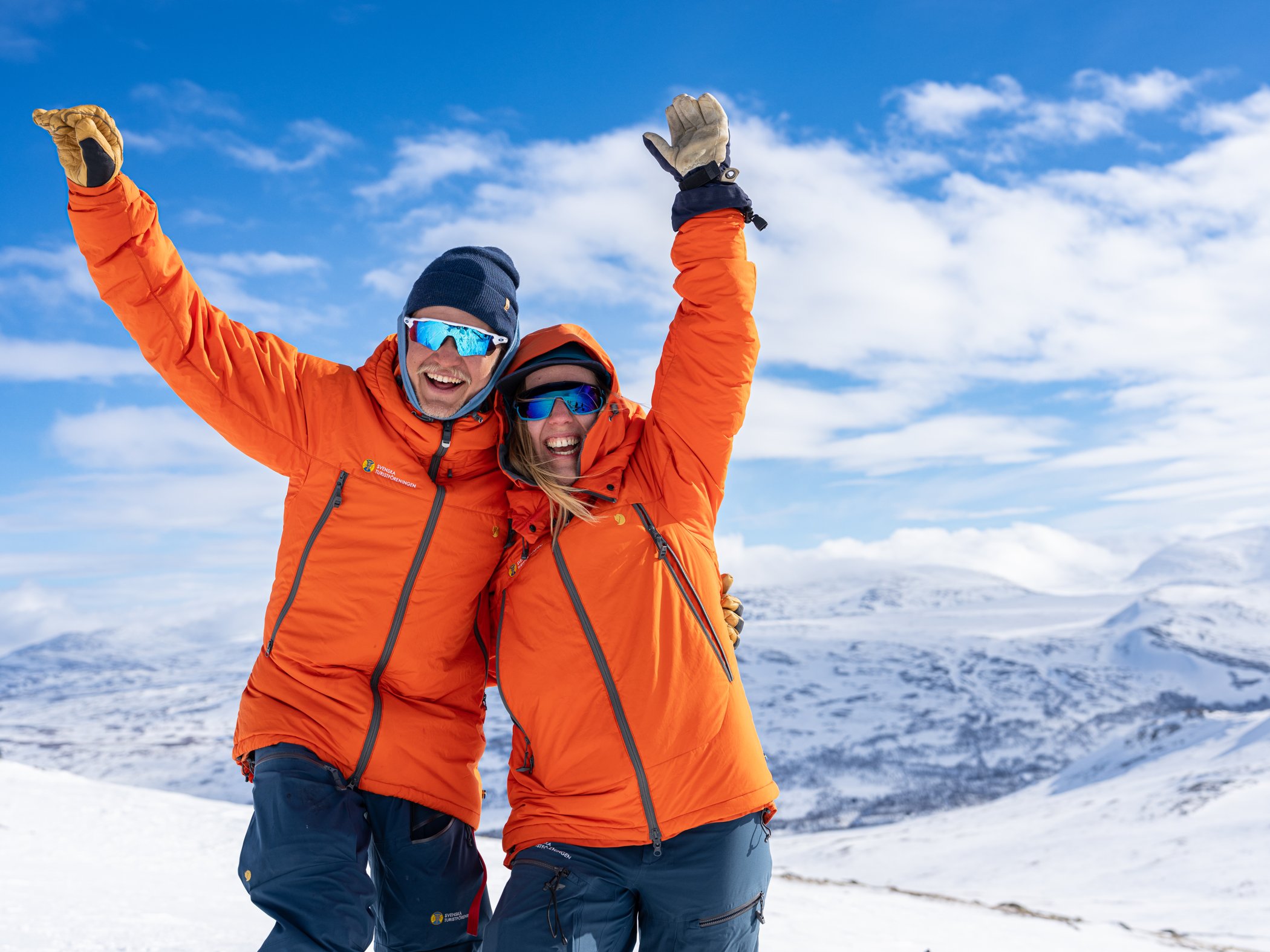 Två personer i orange vinterkläder firar på fjället med snötäckta berg i bakgrunden.
