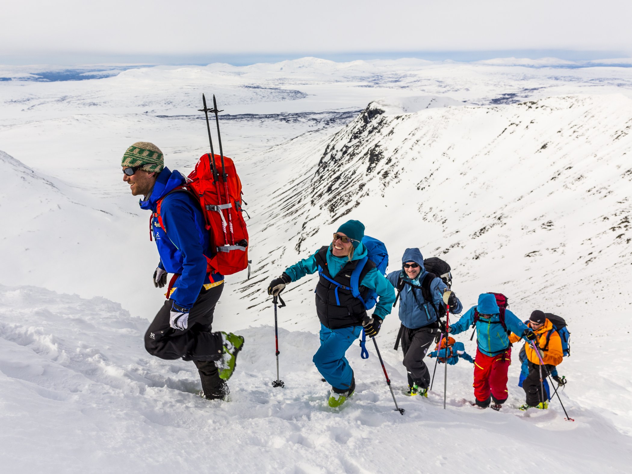 Grupp med toppturåkare som tar sig uppför en snötäckt fjällsida med skidor och stavar.